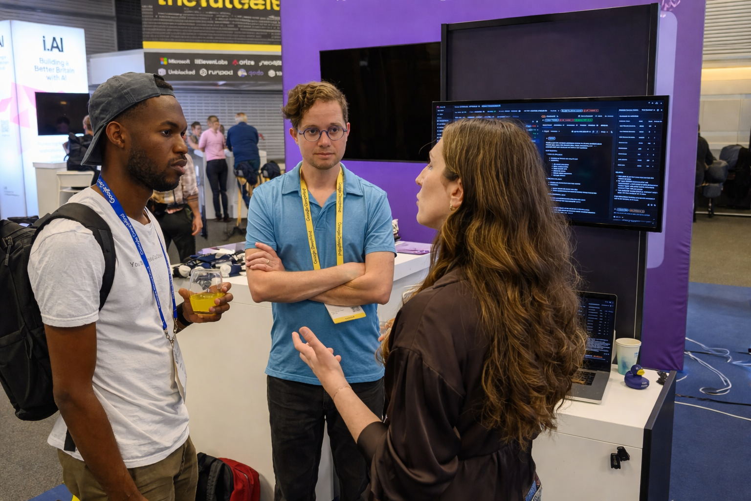 Three developers in discussion at a tech conference booth featuring AI agent observability dashboards, representing collaboration and real-world insights into building production AI systems.